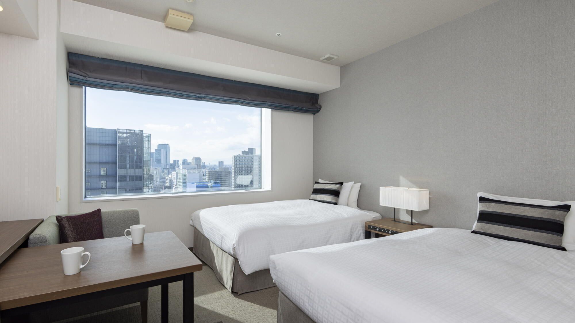 A hotel room with two beds, a desk, a chair, a coffee cup, a lamp, and a view of the city skyline.