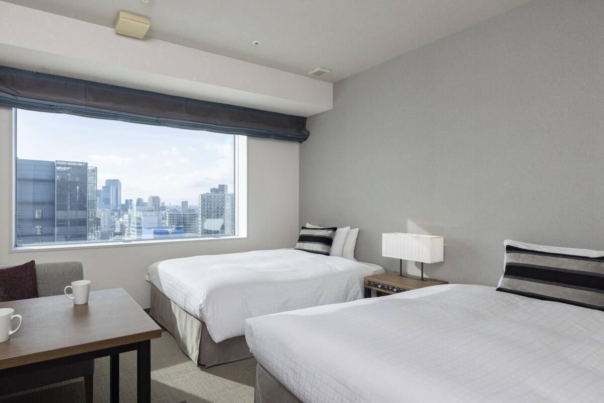 A hotel room with two beds, a desk, a chair, a coffee cup, a lamp, and a view of the city skyline.
