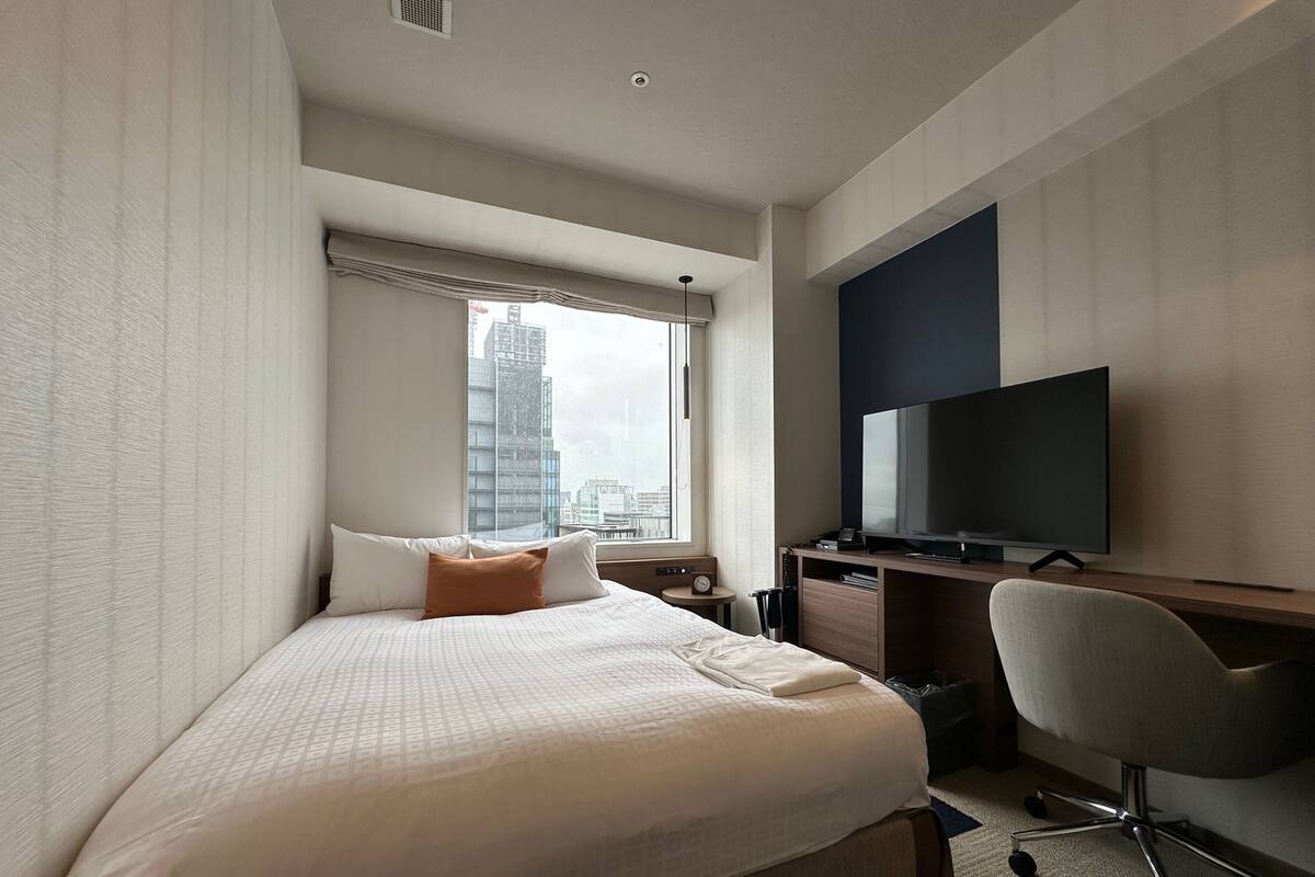A hotel room with a large bed, a desk with a television, and a view of a cityscape through the window.