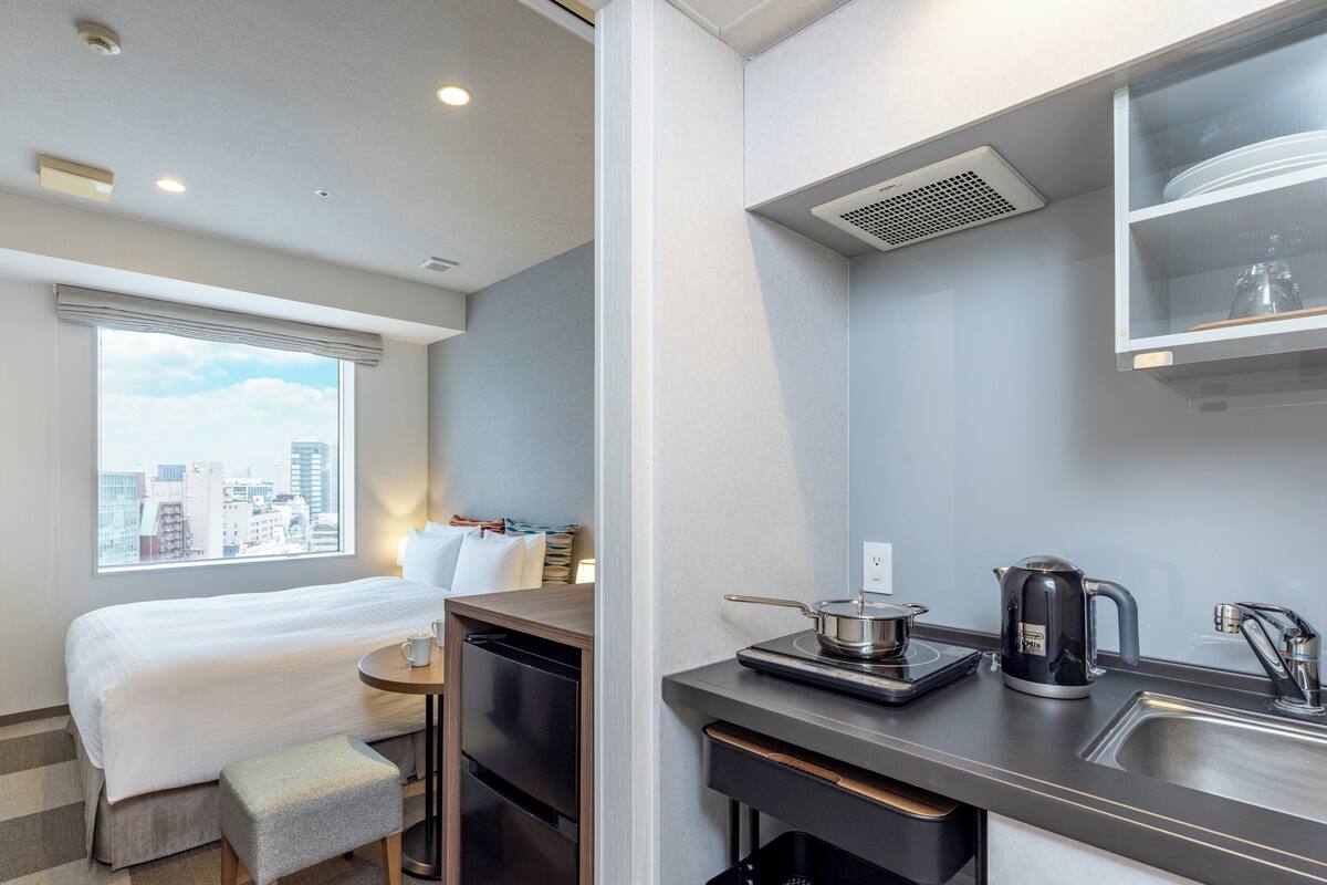 A compact hotel room with a bed, a small table, a kitchenette, and a window with a city view.