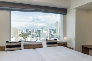 Habitación de hotel con dos camas, un ventanal con vistas a la ciudad, una mesita de noche con lámpara y un cabecero de madera.