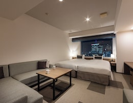 Habitación de hotel moderna con una cama grande, un sofá, una mesita y vistas a un paisaje urbano nocturno.