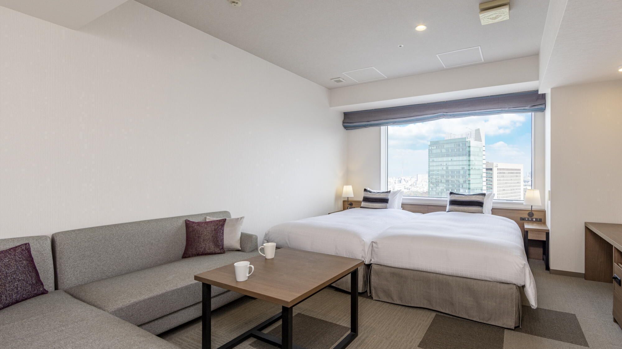 A hotel room with a bed, a sofa, a small table, and a window with a city view.