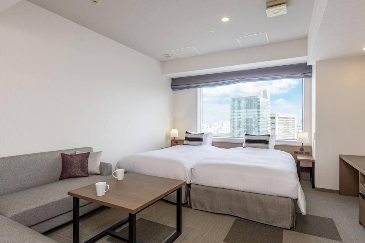 A hotel room with a bed, a sofa, a small table, and a window with a city view.