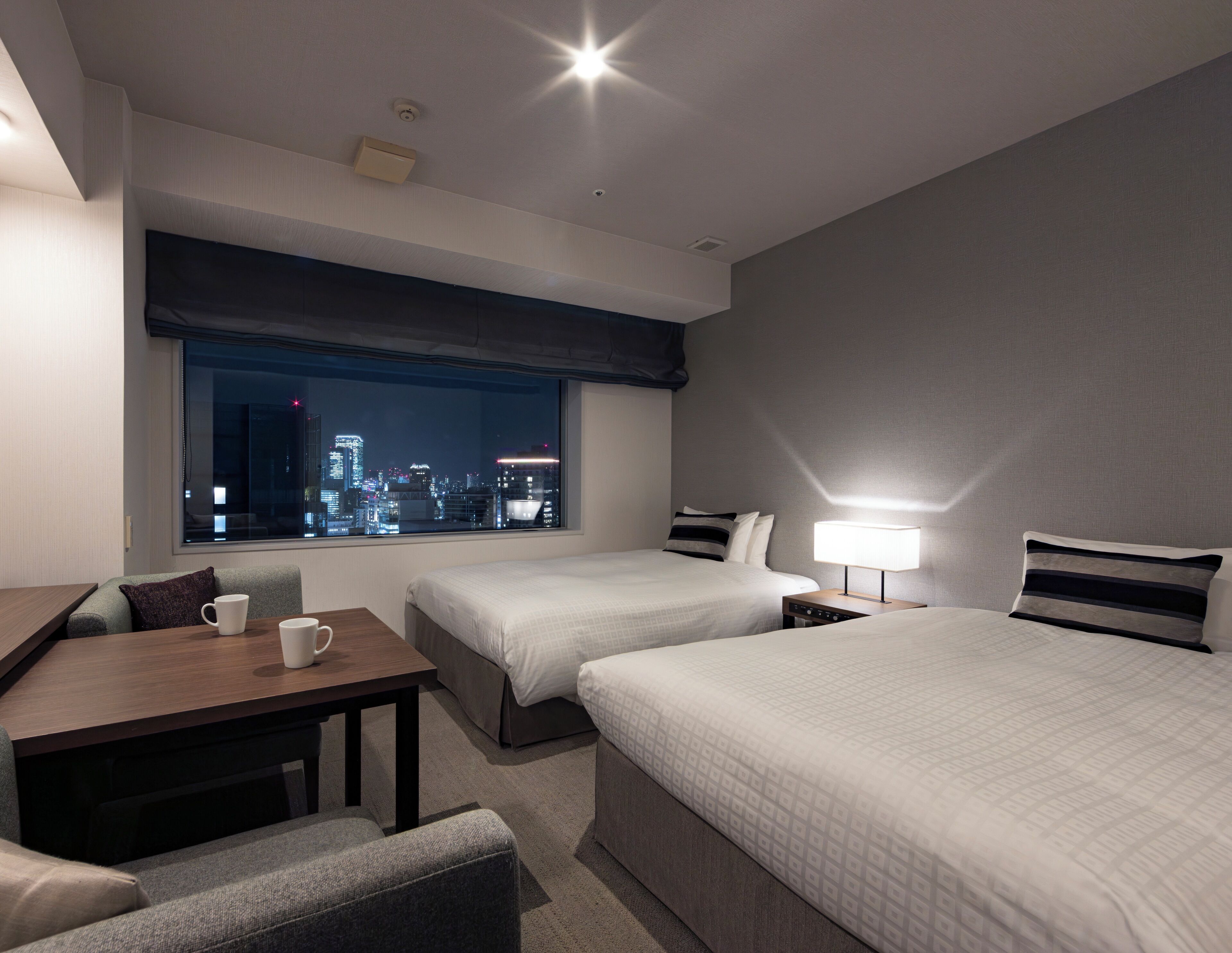 A hotel room with two beds, a desk, a chair, and a view of a cityscape at night.