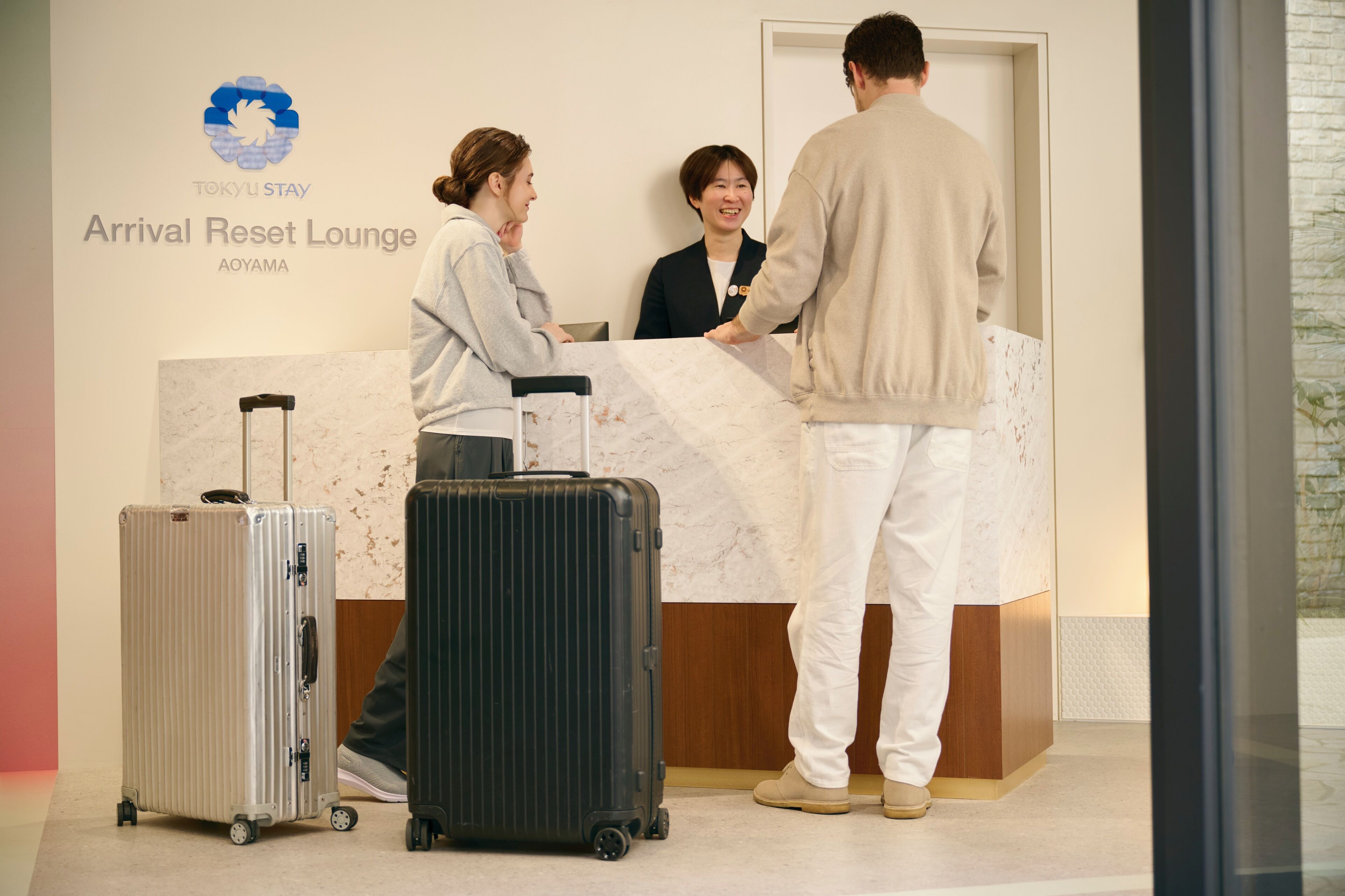 A reception desk with a logo, a counter, and a wall with text. There are two suitcases and a person standing.