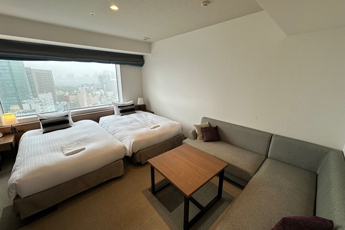 A hotel room with two beds, a sofa, a wooden table, and a view of the city through large windows.