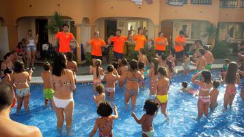 Zona de piscina con gente en traje de baño, algunos adultos con camisetas naranja, y un edificio con balcones al fondo.