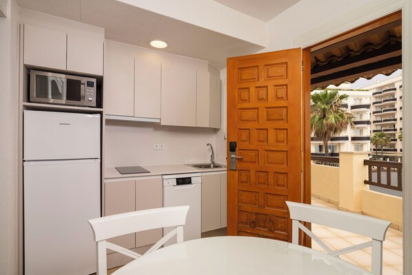 A compact kitchen with a microwave, refrigerator, and dishwasher. A wooden door leads to an outdoor balcony with a view of a residential building.