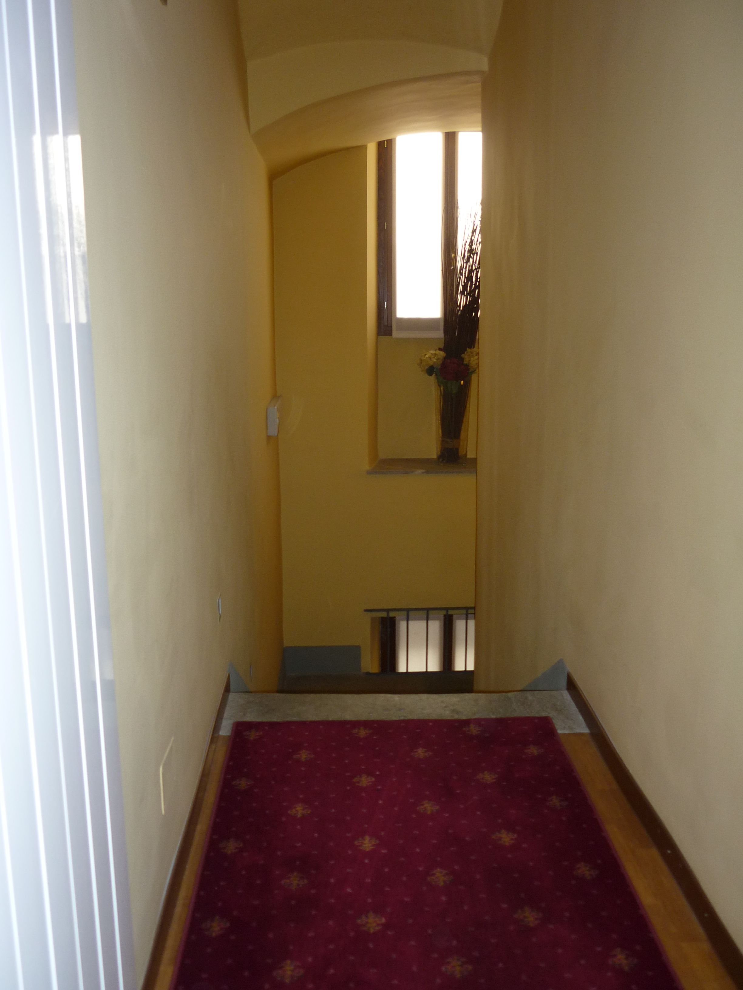 A narrow hallway with a red carpet, a window at the end, and a small balcony with a railing.