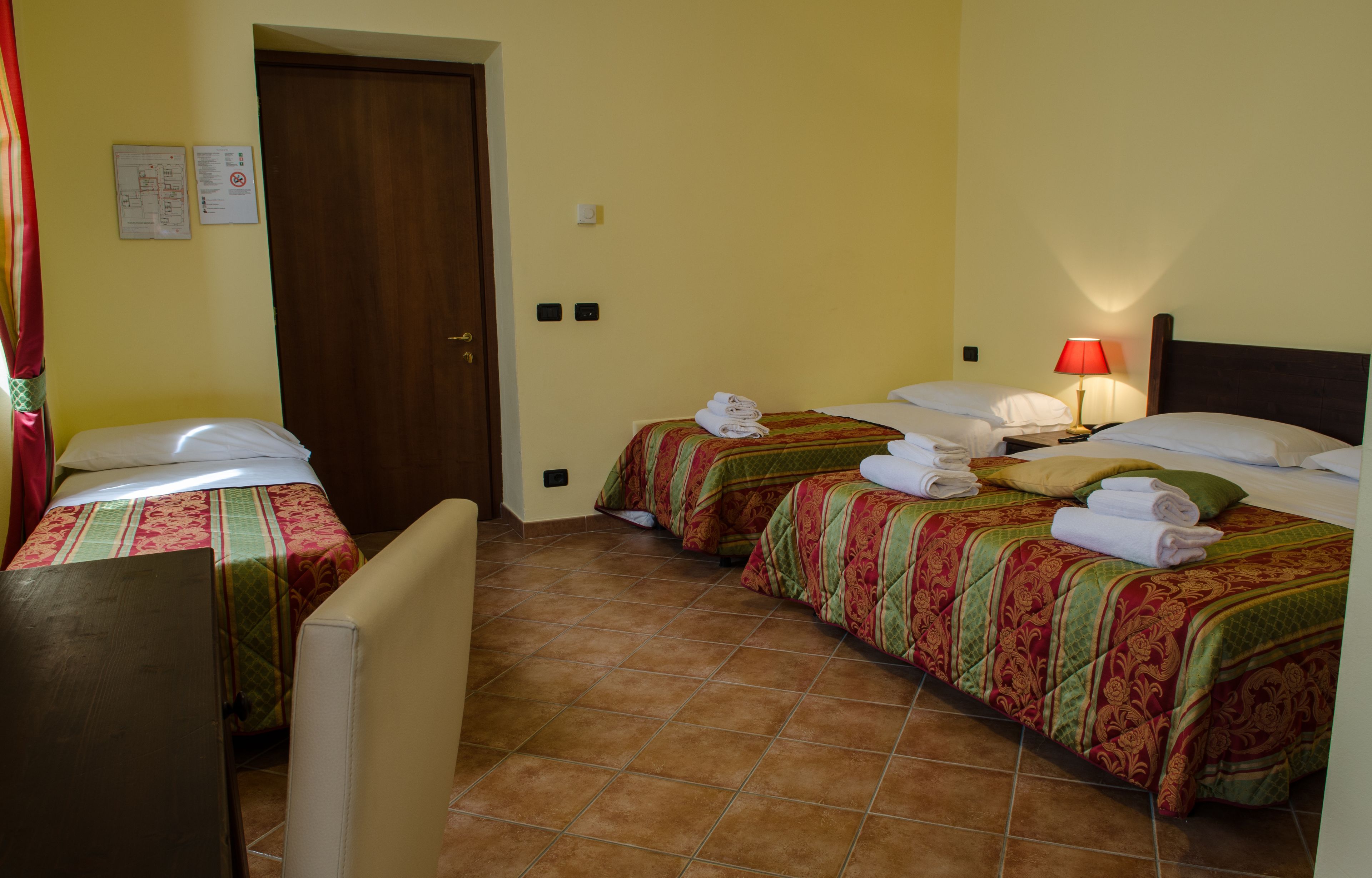A hotel room with two single beds, each with a green and red patterned bedspread and white pillows. There is a wooden door, a small table, and a wall-mounted light fixture.
