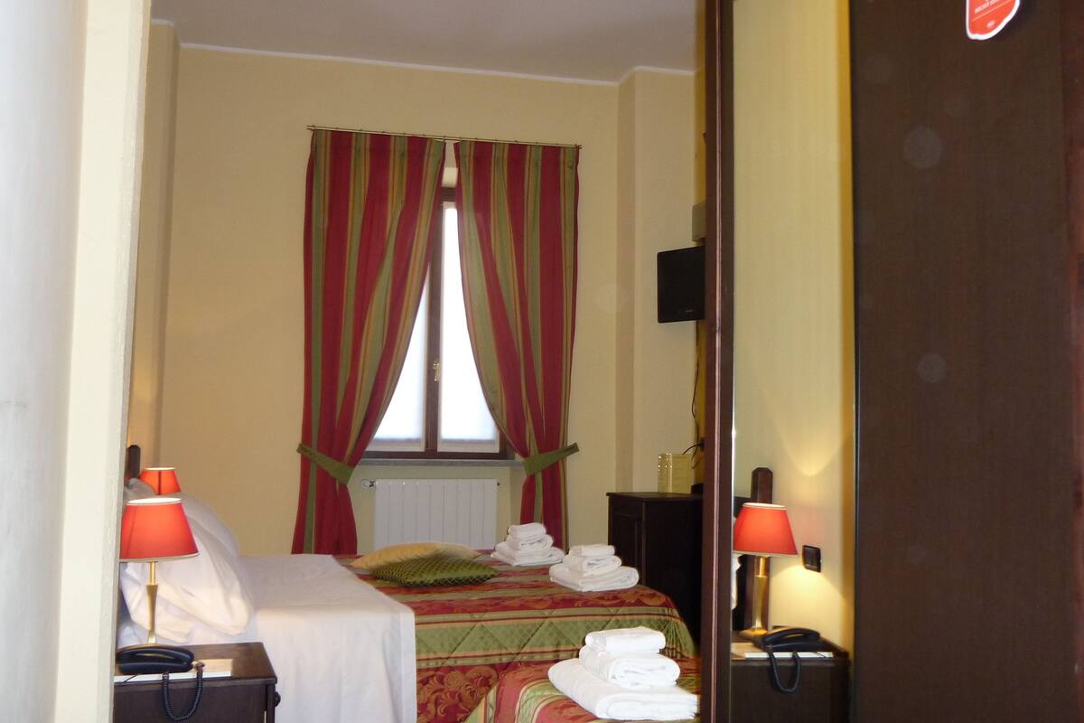 A hotel room with a bed, two bedside lamps, a mirror, a window with curtains, and a radiator.