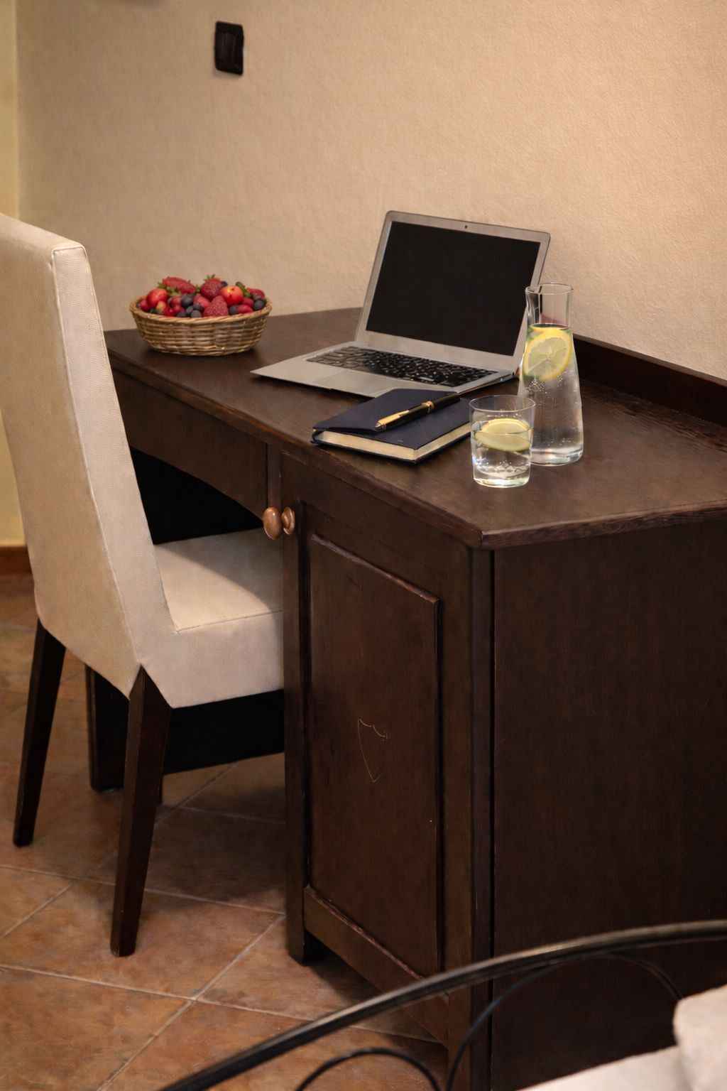 Ein Holztisch mit einem Laptop, einem Notizbuch, einem Stift, einem Glas Wasser mit Zitrone und einer Fruchtschale.