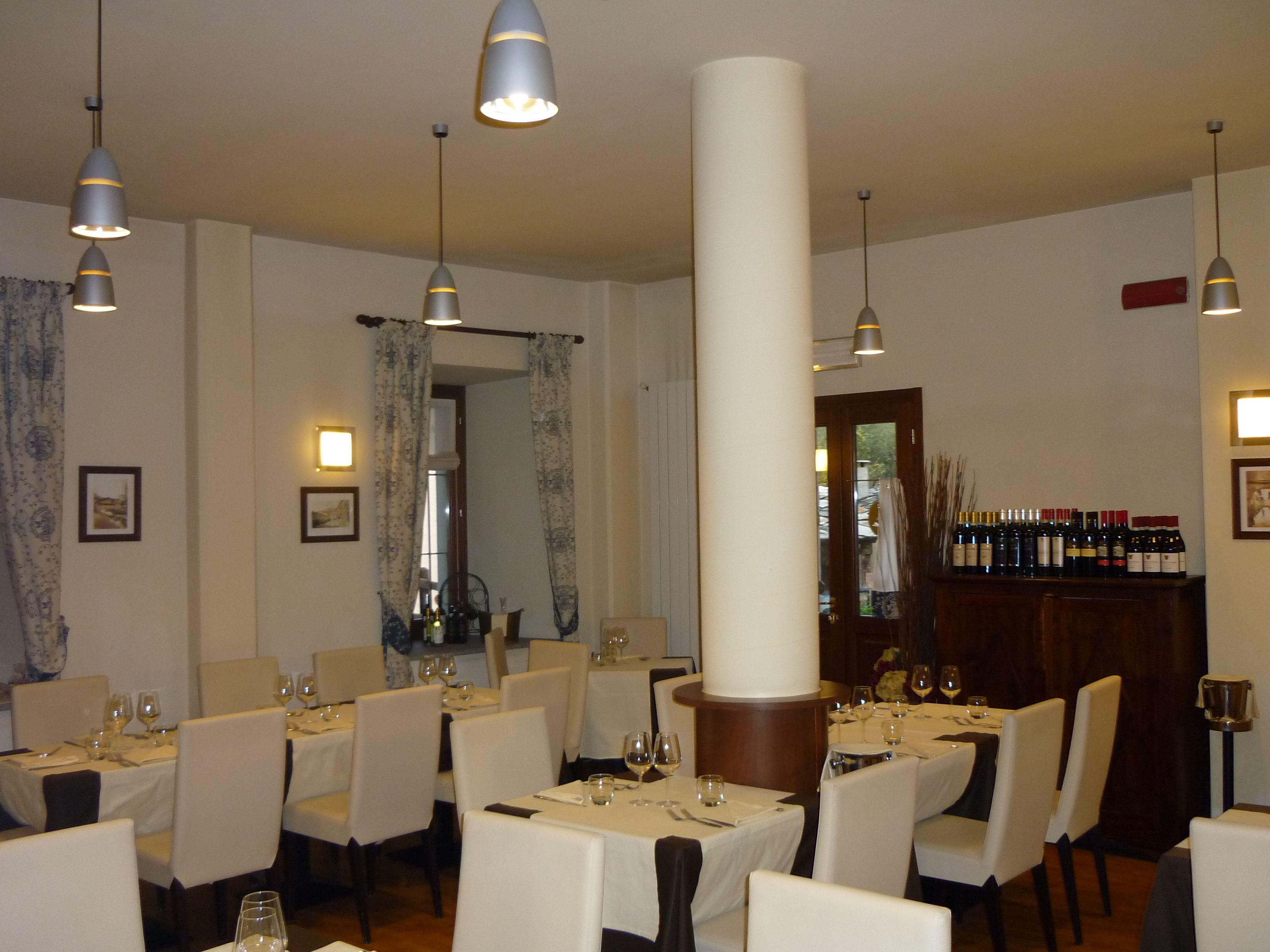 A restaurant interior with multiple tables and chairs, pendant lighting, and a wine display.