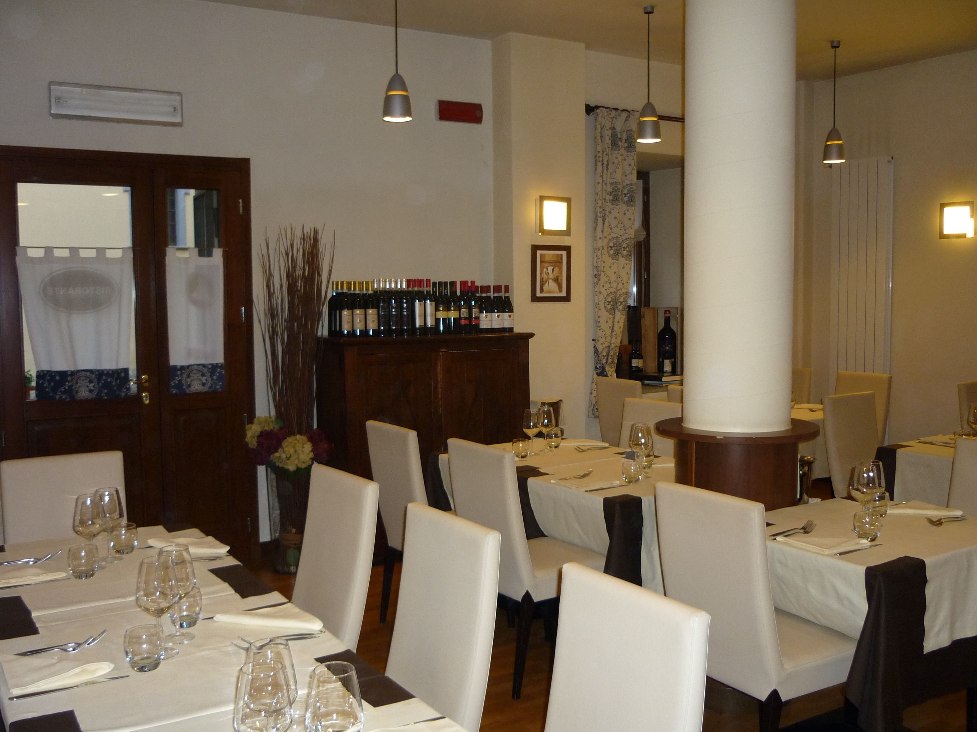 Un comedor con mesas dispuestas para cuatro, cada una con copas de vino, cubiertos y un jarrón de flores. Hay botellas de vino en una estantería, y la habitación tiene un diseño clásico con muebles de madera e iluminación colgante.