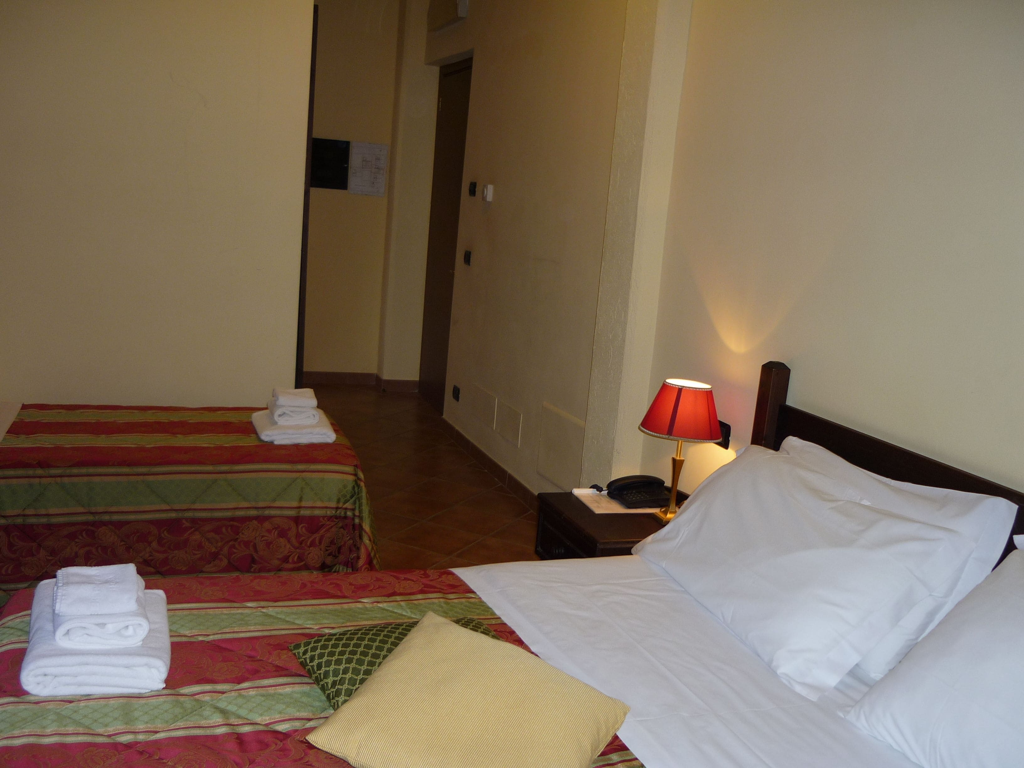 A hotel room with two beds, a nightstand with a lamp, and a hallway leading to another room.