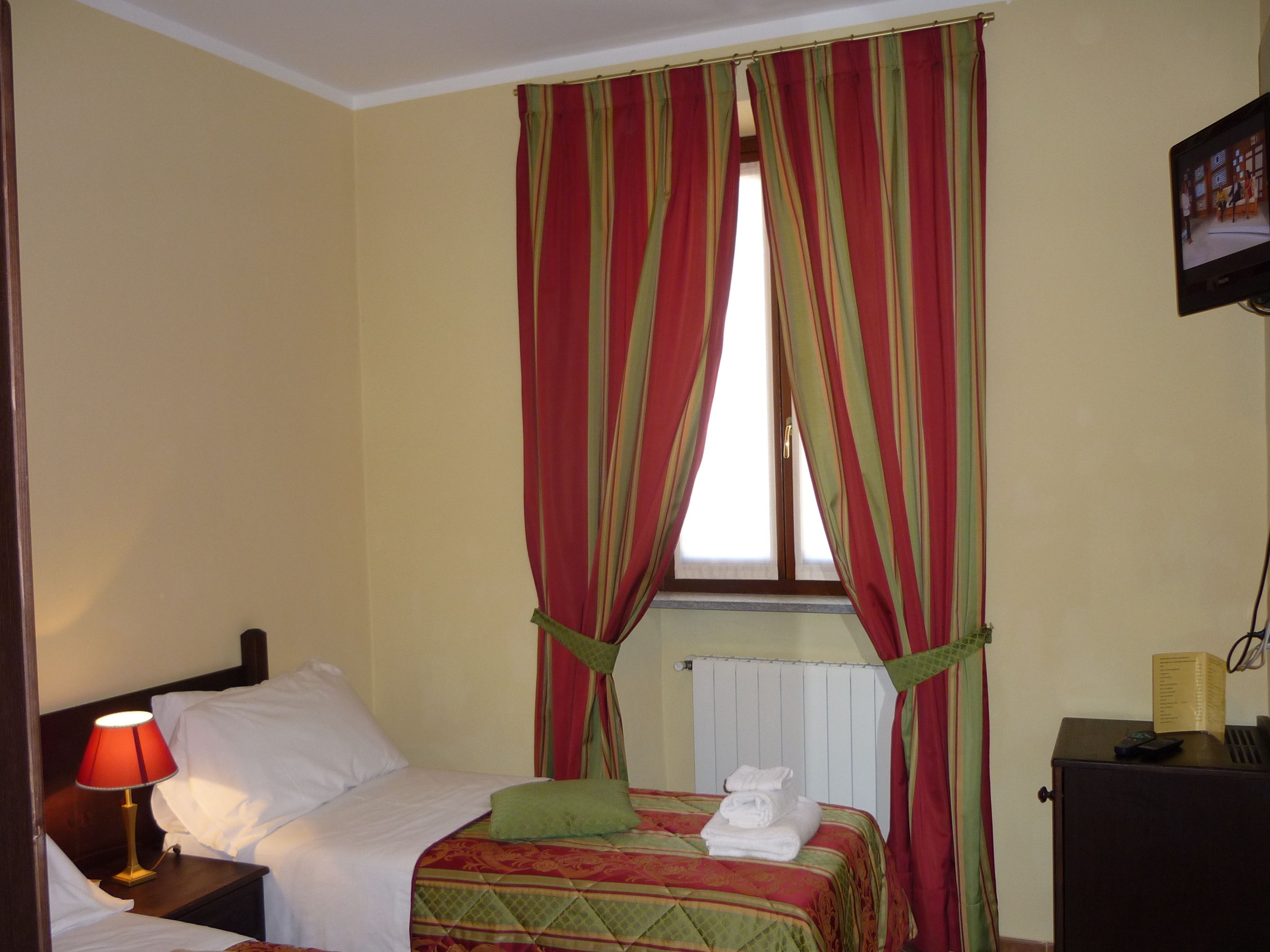 A hotel room with a bed, a nightstand with a lamp, a window with curtains, a radiator, and a television on the wall.