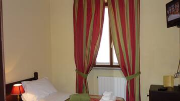 A hotel room with a bed, a nightstand with a lamp, a window with curtains, a radiator, and a television on the wall.