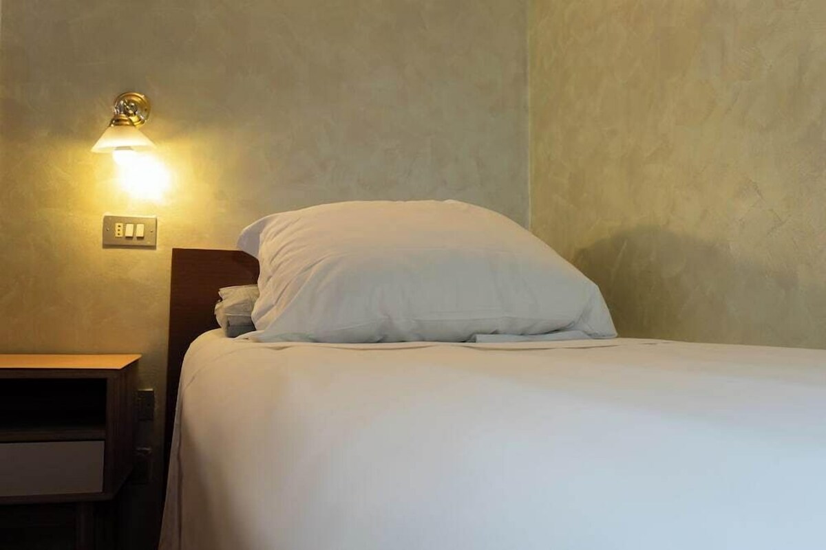 Un letto singolo con biancheria bianca, un comodino con una lampada e una presa a muro.