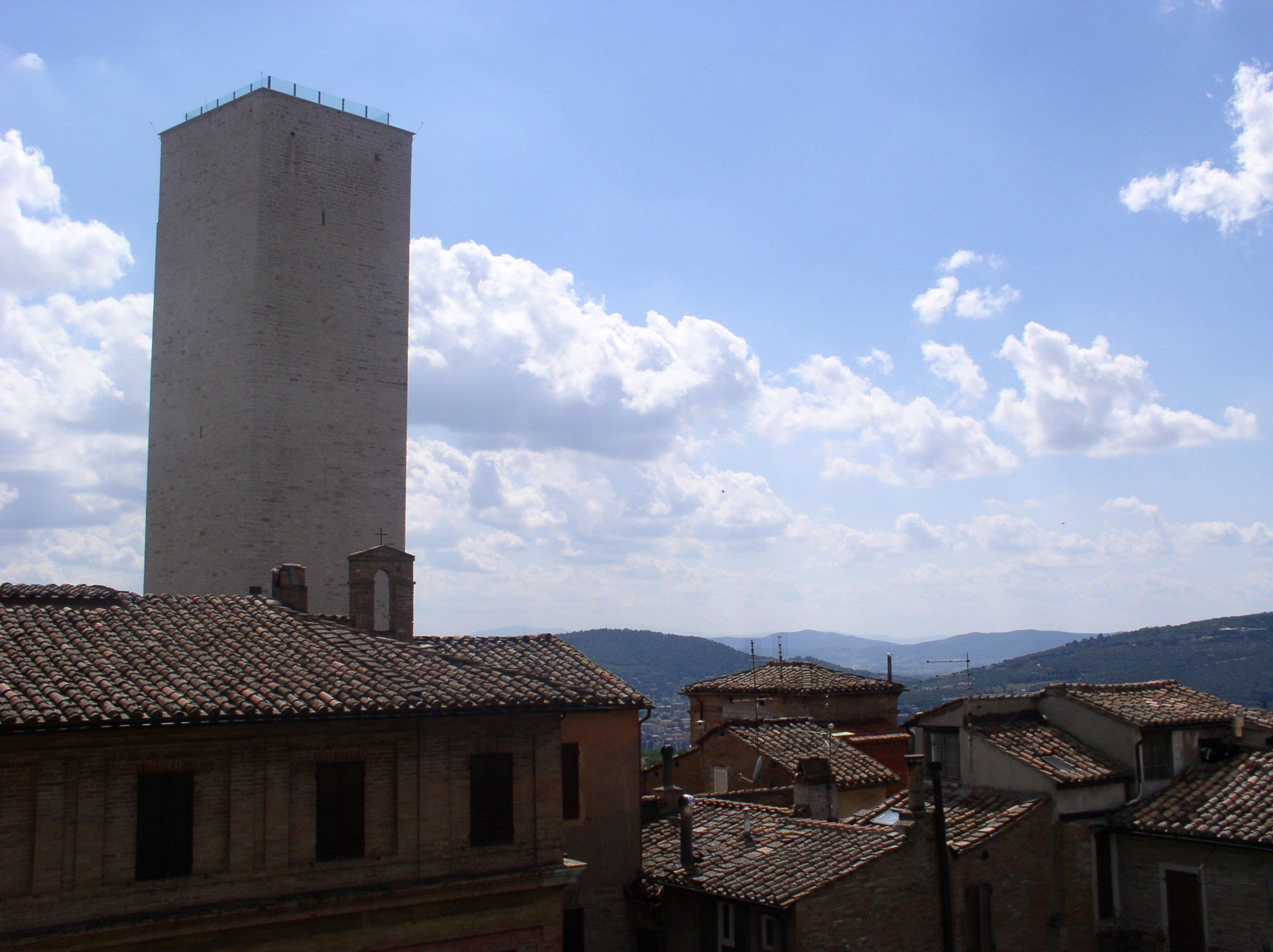 A tall, rectangular tower with a cross at the top, surrounded by traditional buildings with terracotta roofs.