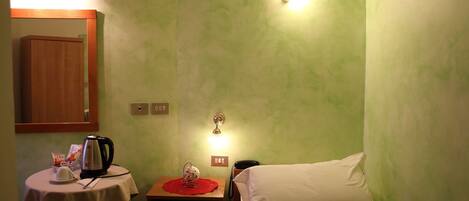 A small hotel room with a single bed, a bedside table with a kettle, a telephone, and a small table with a red placemat.