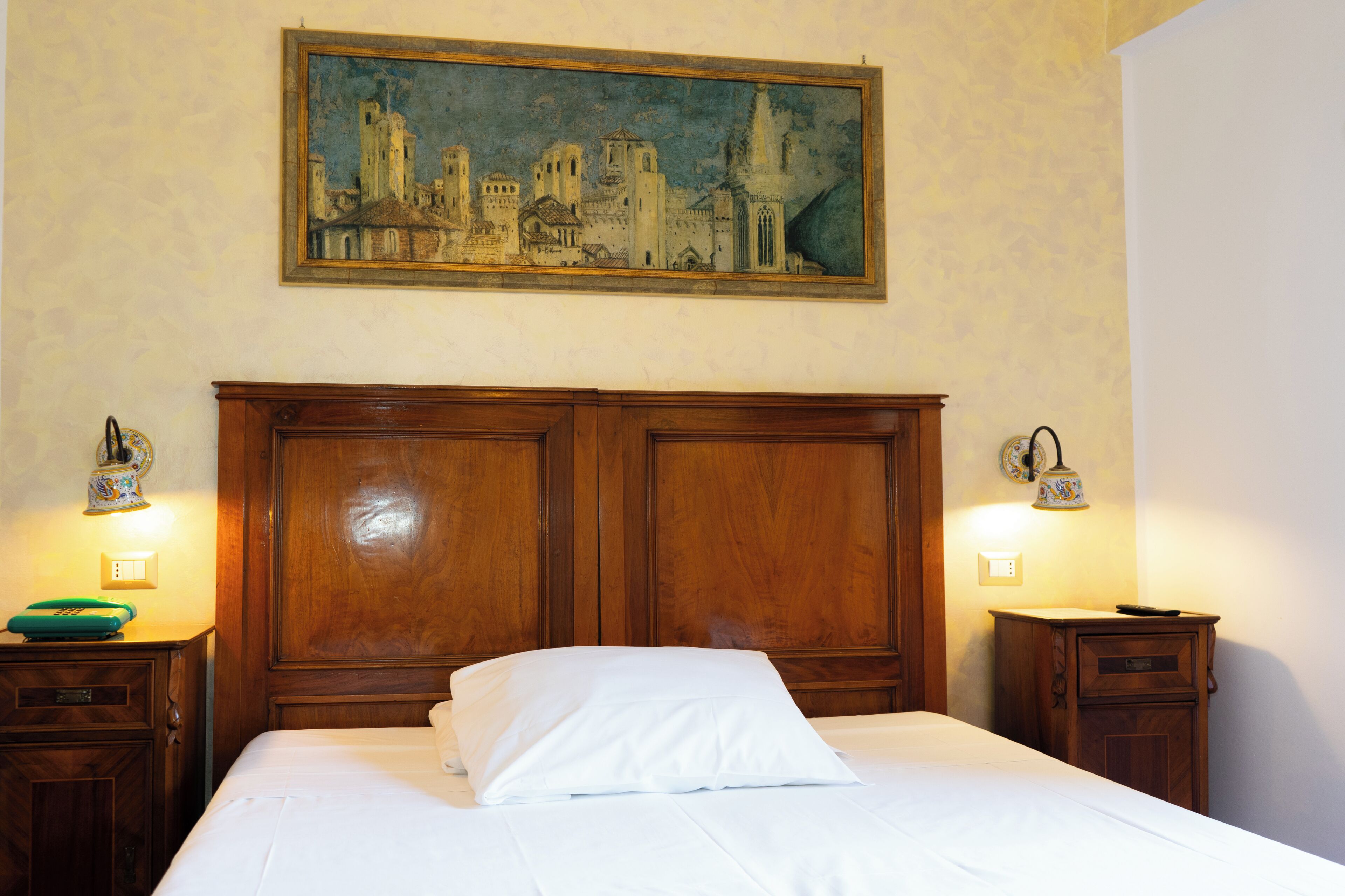 Ein Hotelzimmer mit hohem, hölzernem Kopfteil, einem Stadtbildgemälde an der Wand und zwei Nachttischen mit Lampen.