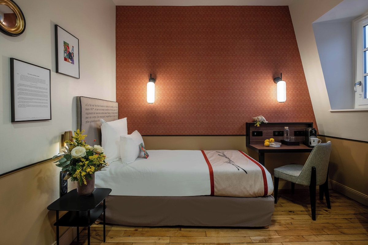 A bedroom with a bed, a desk, a chair, a small table with a vase of flowers, and two wall-mounted lights.
