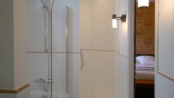 A bathroom with a shower, a toilet, and a sink. There is a bed visible in the adjacent room.