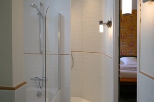 A bathroom with a shower, a toilet, and a sink. There is a bed visible in the adjacent room.