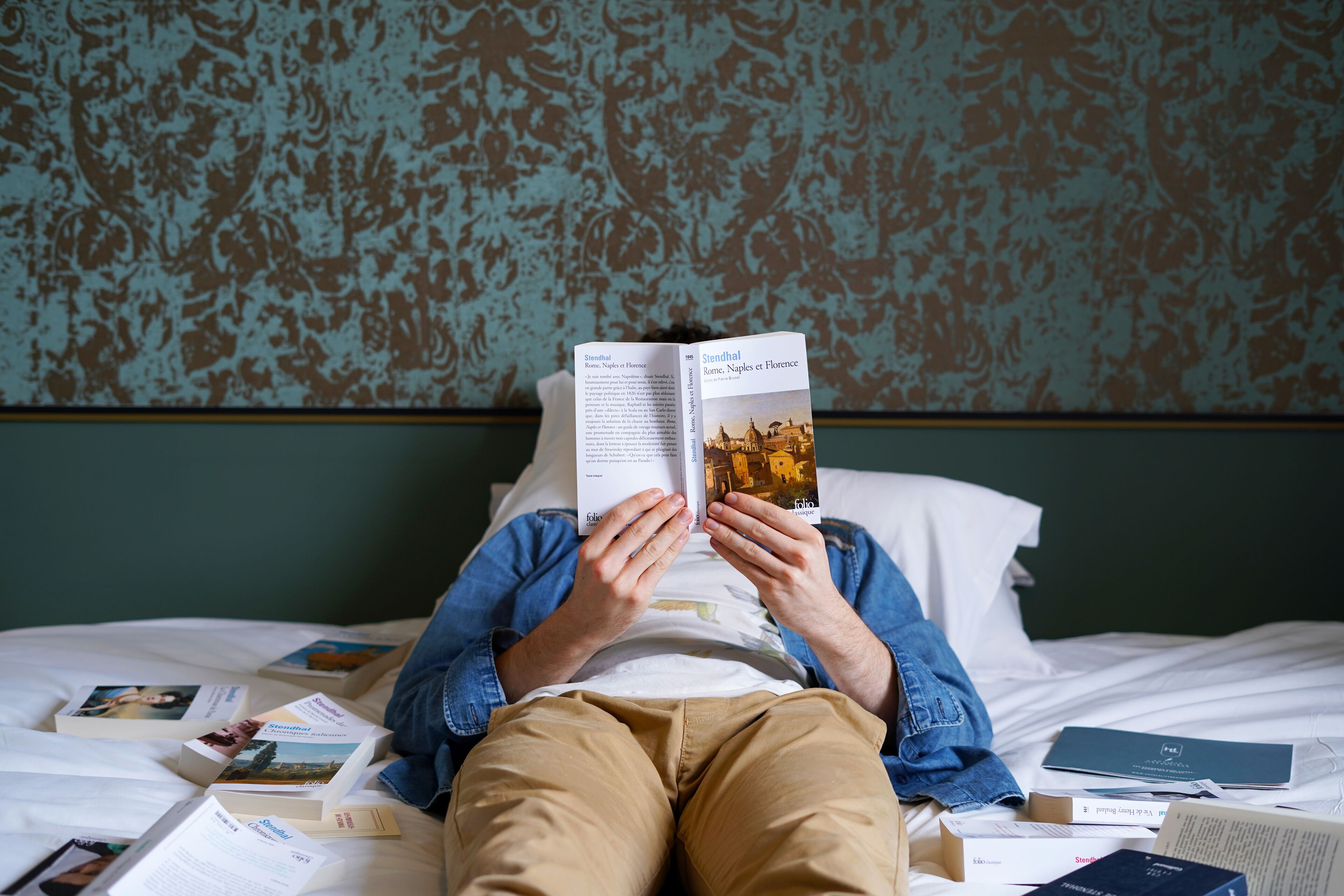 A person lying on a bed with a book open, surrounded by other books.