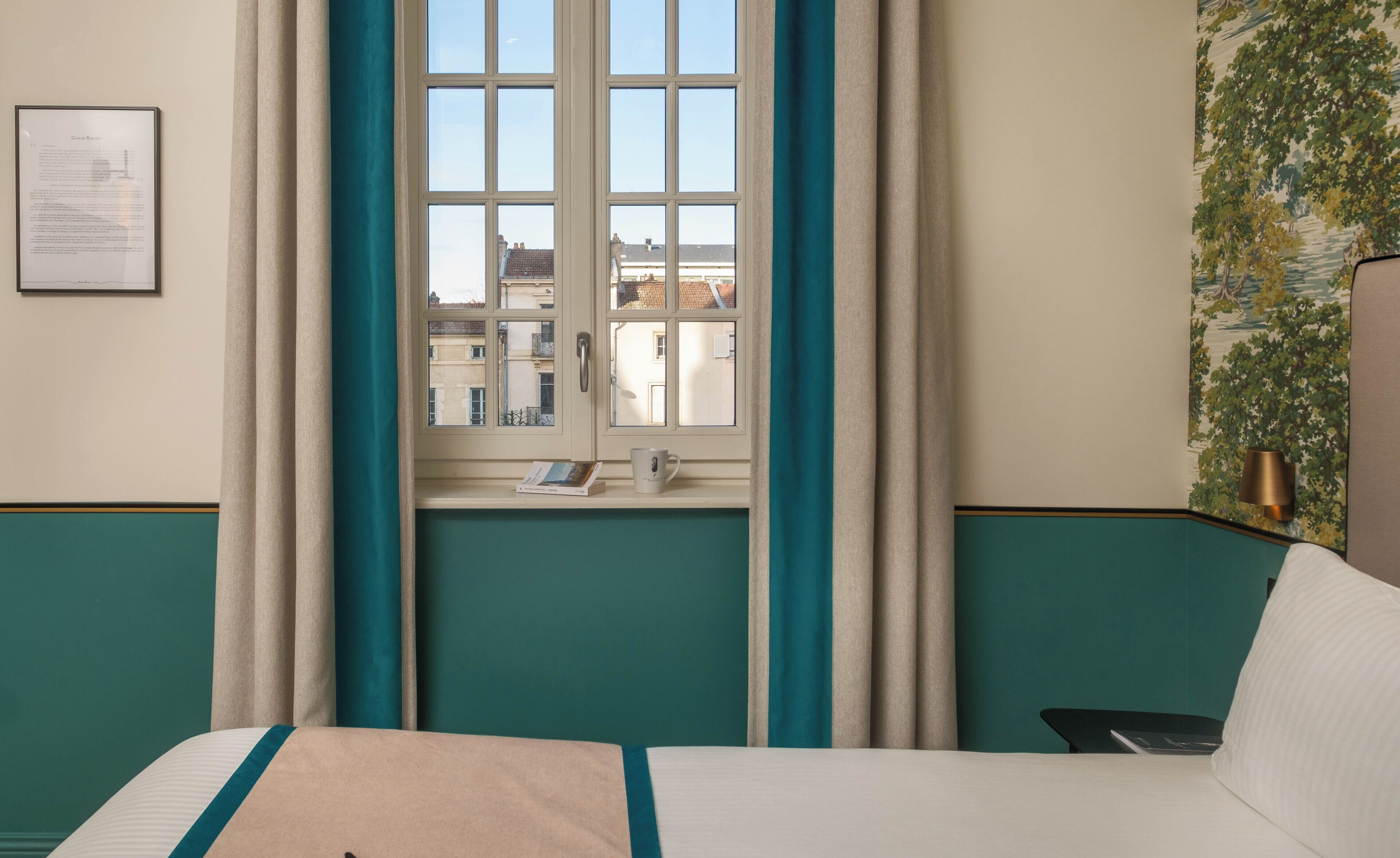 A hotel room with a bed, a window with curtains, a view of buildings outside, and a wall with a mural.
