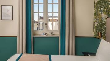 A hotel room with a bed, a window with curtains, a view of buildings outside, and a wall with a mural.