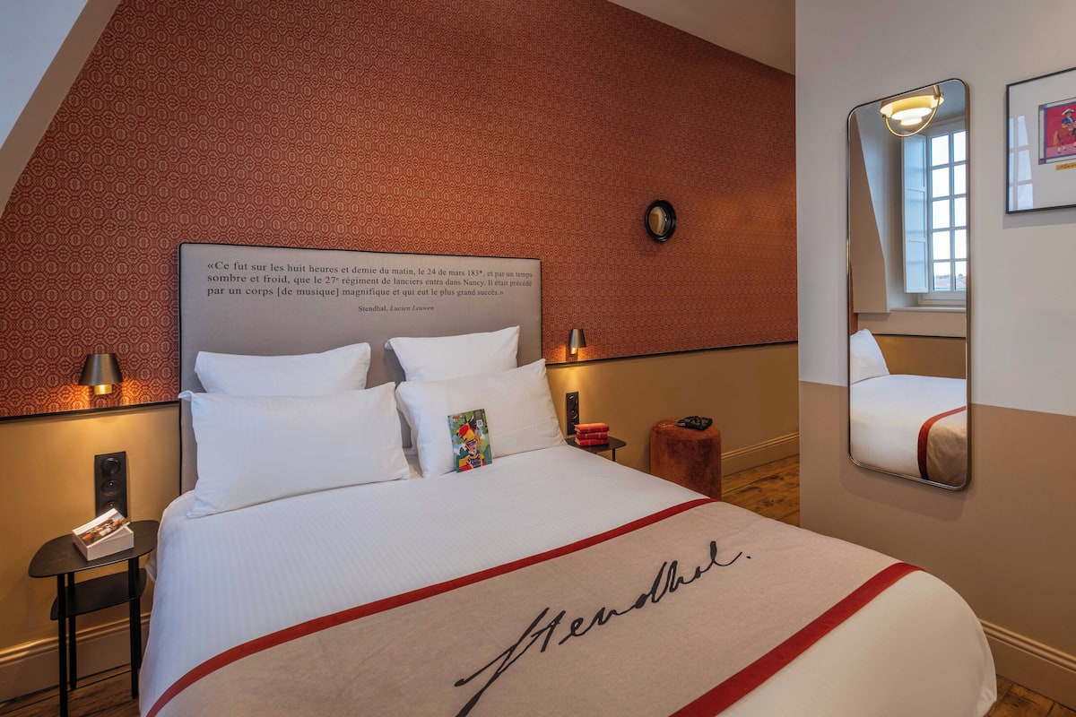 A hotel room with a large bed, a headboard with a quote, a bedside table with a lamp, a small nightstand, a mirror, and a framed picture on the wall.