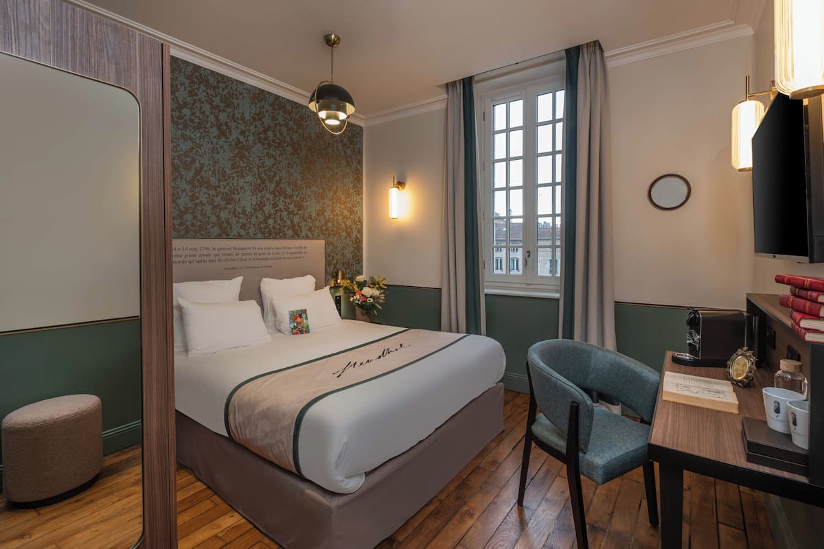 A hotel room with a bed, a desk with a chair, a window with curtains, a wall clock, and a bookshelf with books.