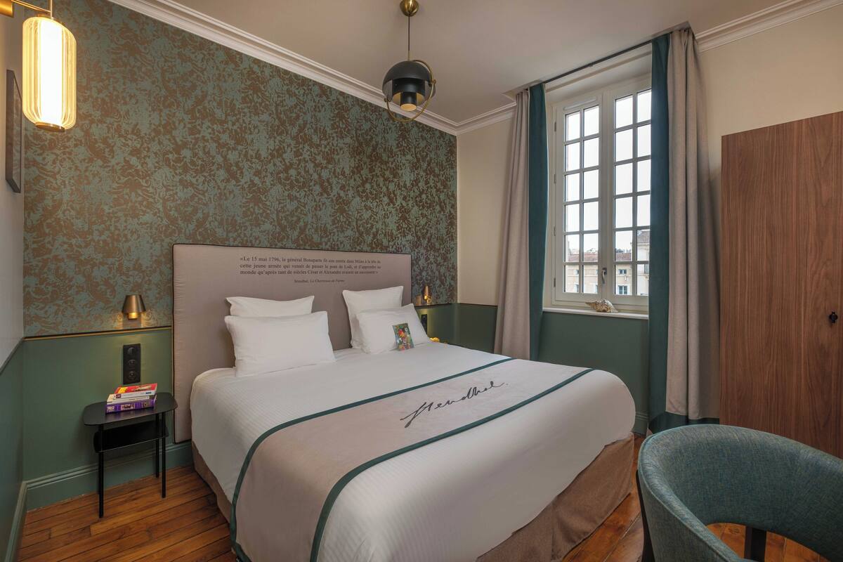 A hotel room with a large bed, a bedside table with books, a lamp, and a view of the city through a window.