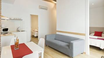 A modern room with a grey sofa, a dining table with a red table runner, a kitchen area with a microwave, and a bed with a red pillow.