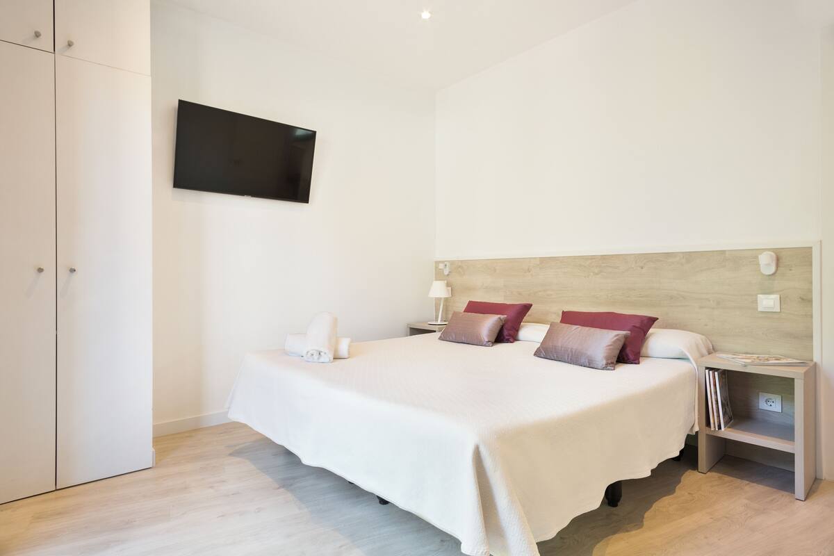 A neatly arranged bedroom with a bed, bedside tables, a TV mounted on the wall, and a wooden headboard.