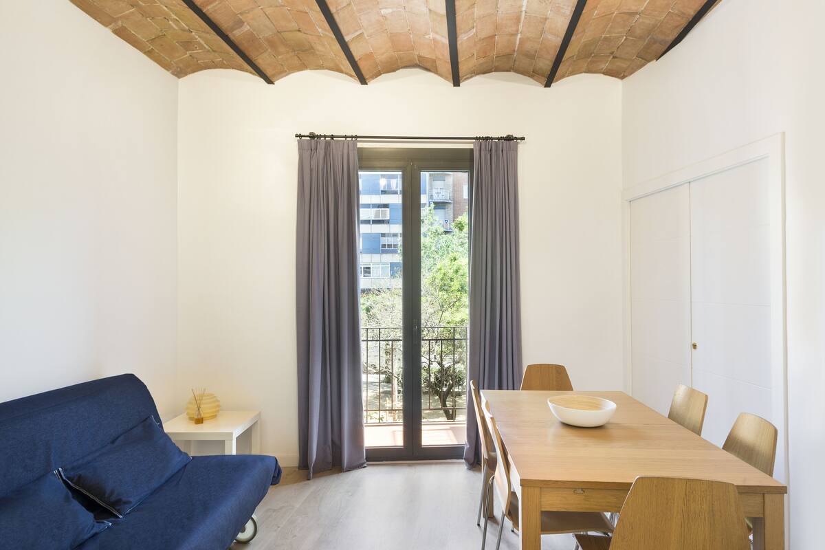 A room with a dining table, chairs, a sofa, and a sliding glass door leading to a balcony.