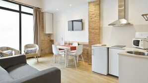 A compact kitchen with a microwave, a small refrigerator, and a sink. There is a dining area with a table and chairs, and a sofa. A television is mounted on the wall above the dining area. - BcnStop Parc Güell (Barcelona)