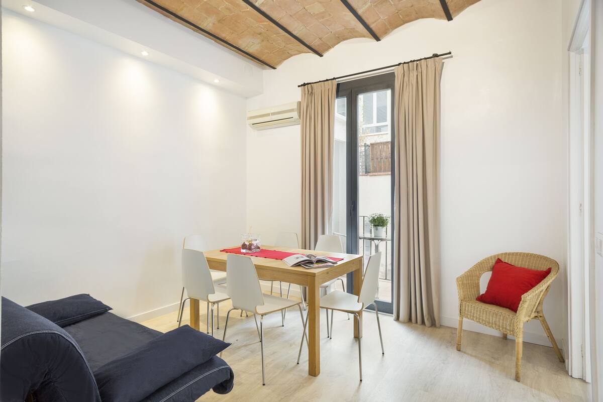 A modern living room with a dining table, chairs, a sofa, and a wicker chair with a red cushion.