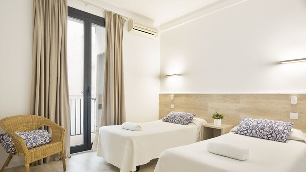 A hotel room with two beds, a wicker chair, a small table, and a potted plant. - BcnStop Parc Güell (Barcelona)