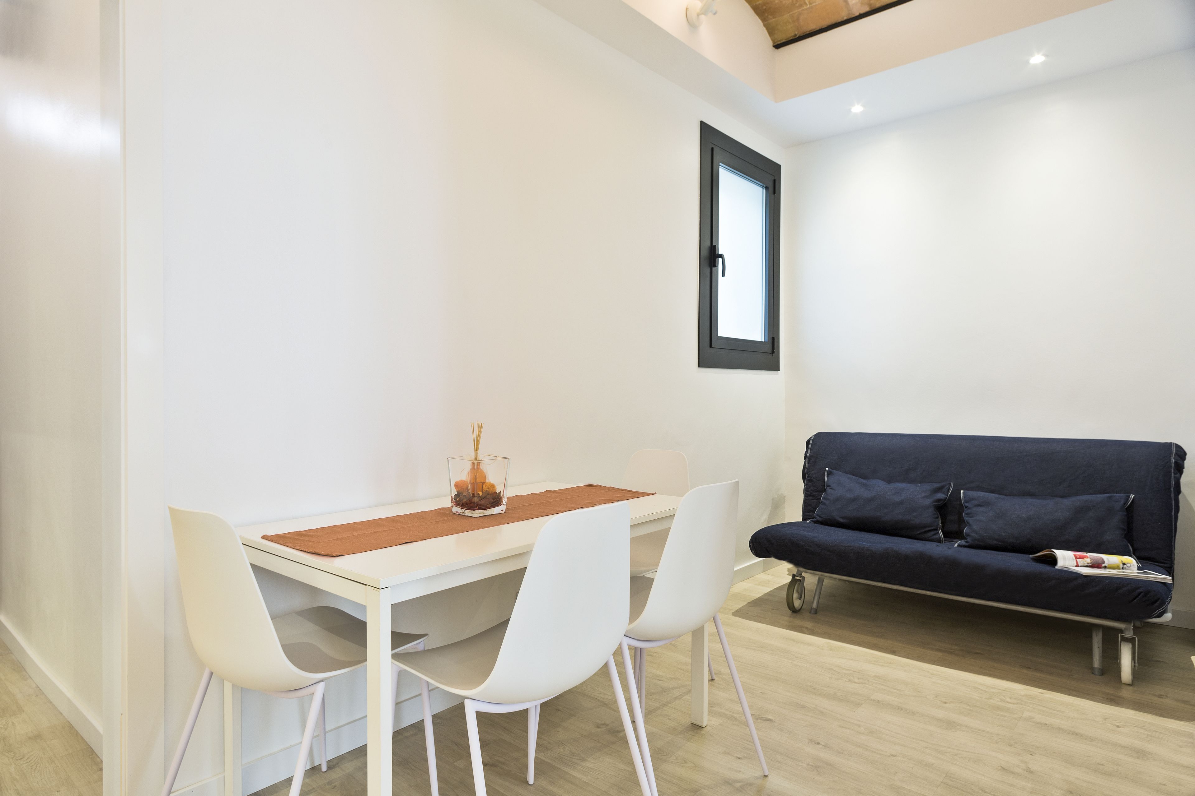 A modern room with a white table, four white chairs, a dark blue sofa, and a window with a black frame.