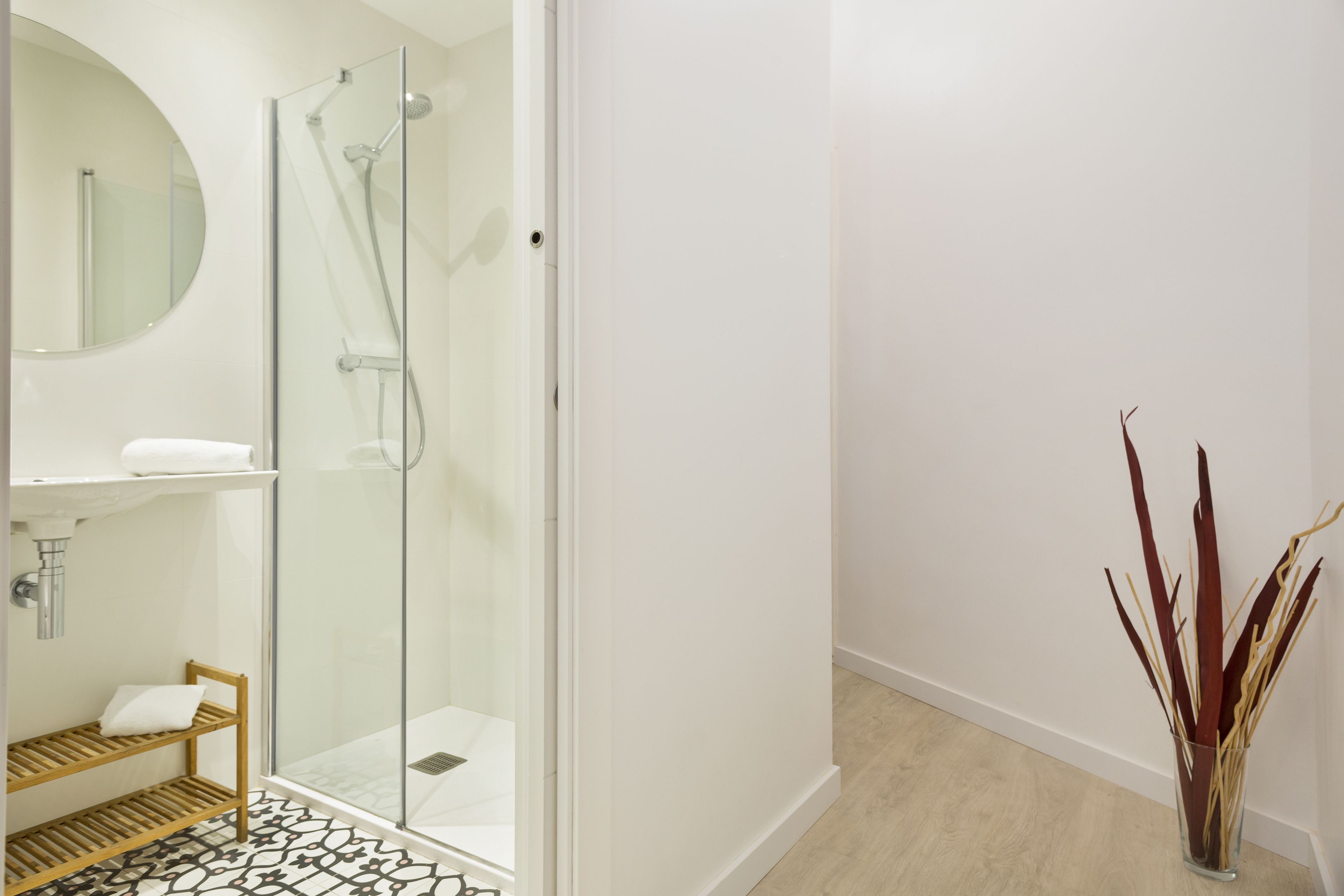 A modern bathroom with a glass-enclosed shower, a round mirror, a wooden shelf, and a vase with decorative plants.