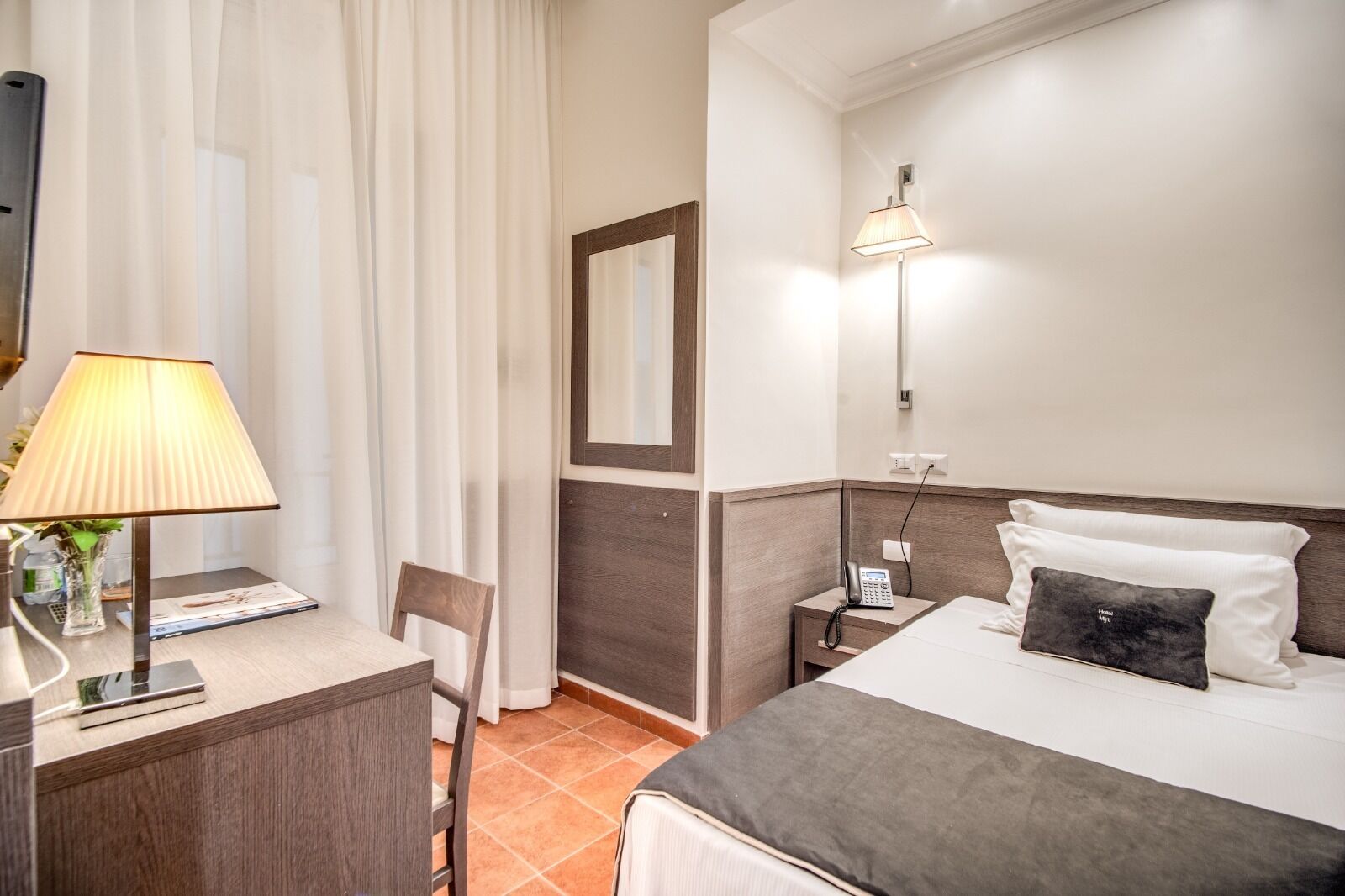 A hotel room with a bed, bedside table, lamp, chair, and a mirror.