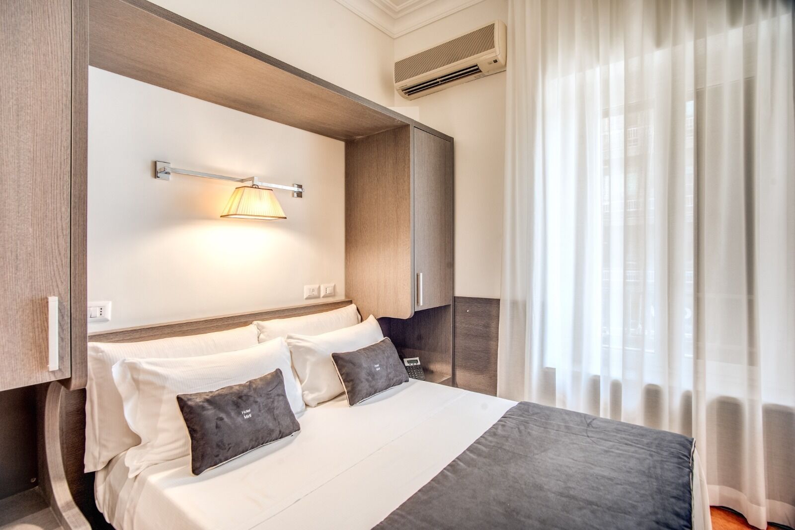 Ein Hotelzimmer mit Bett, Nachttisch, Lampe, Klimaanlage und einem Fenster mit Vorhängen.