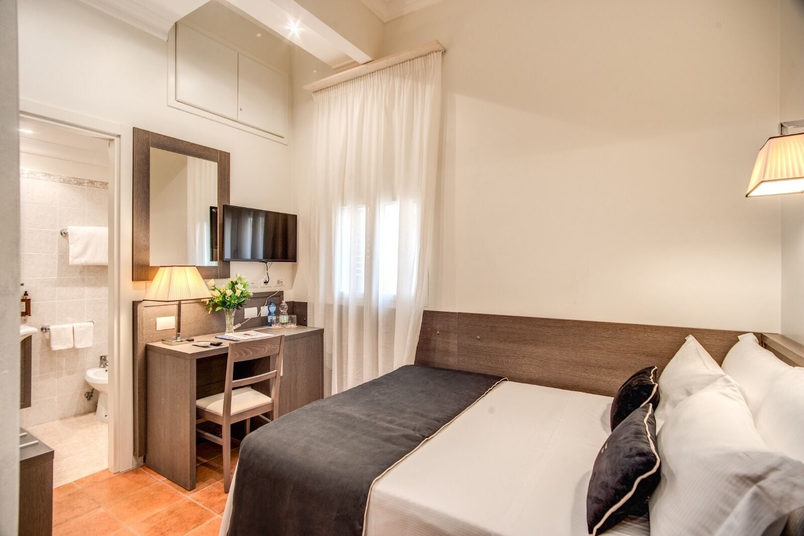 A hotel room with a bed, a desk, a chair, a TV, a mirror, a window with curtains, and a bathroom with a toilet and sink.