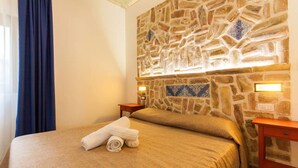 Habitación de hotel con pared de piedra, cama con cubrecama color beige, dos mesitas de noche con lámparas y una cortina azul.