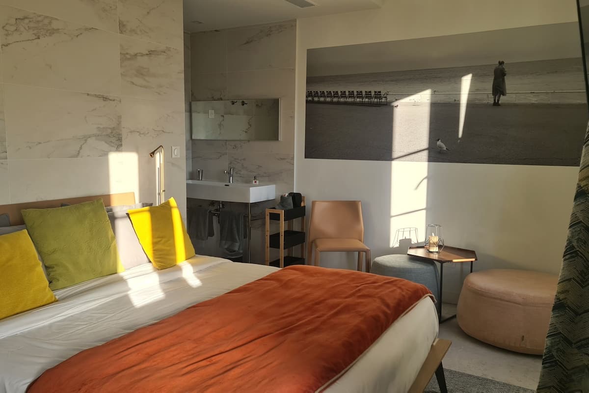 A modern hotel room with a large bed, a sitting area, a small table, a chair, a mirror, and a view of the beach.