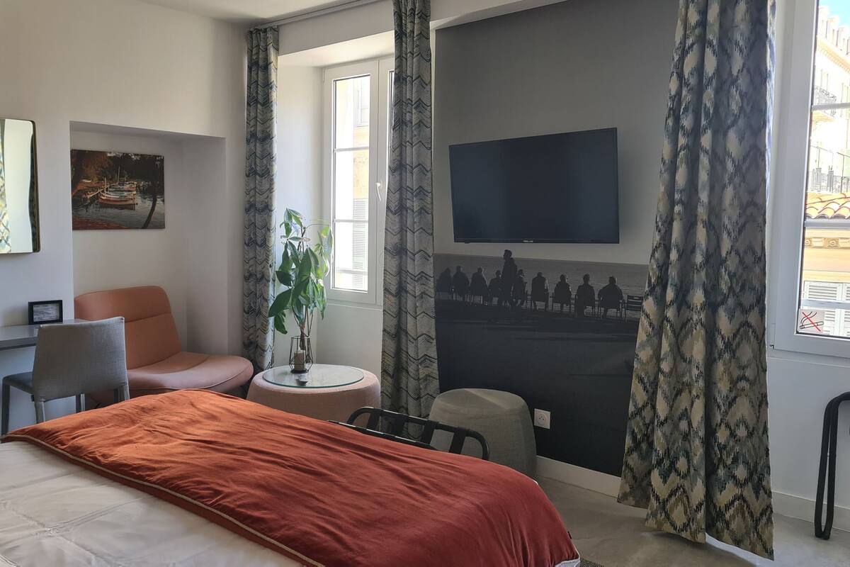 A hotel room with a bed, a desk with a chair, a TV mounted on the wall, and a window with curtains.