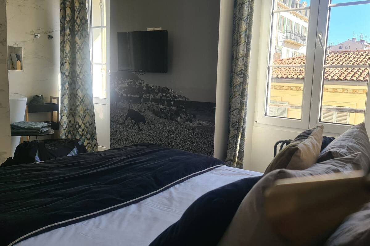 A hotel room with a bed, a TV mounted on the wall, and a window with a view of buildings.